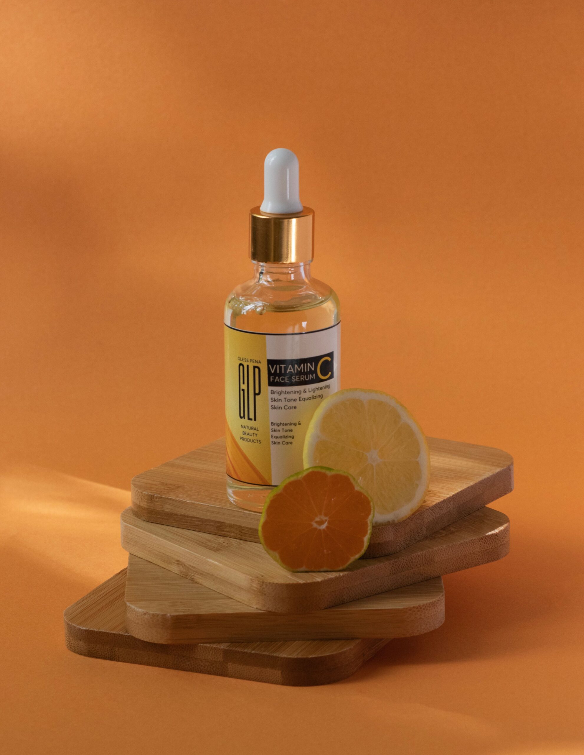 Vitamin C serum bottle with lemon and orange slices on wooden coasters against orange background.